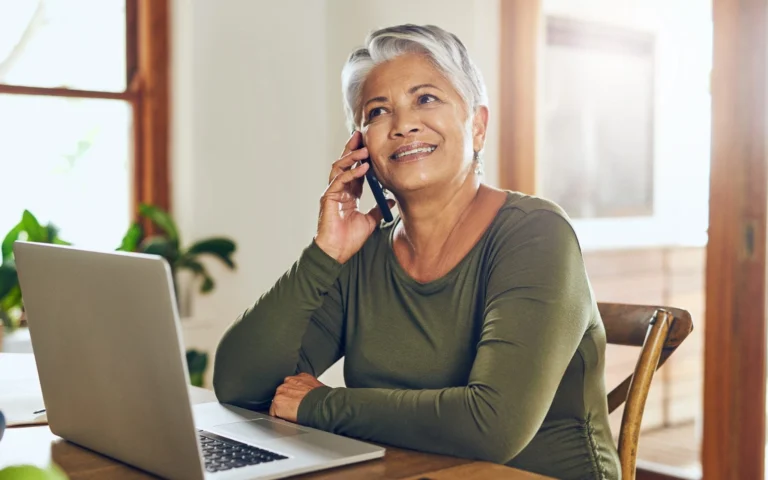 smiling older woman sitting at kitchen table on phone and computer