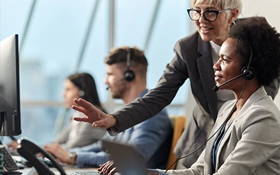 Senior manager coaching a health advisor wearing a headset at a contact center workstation with colleagues in background