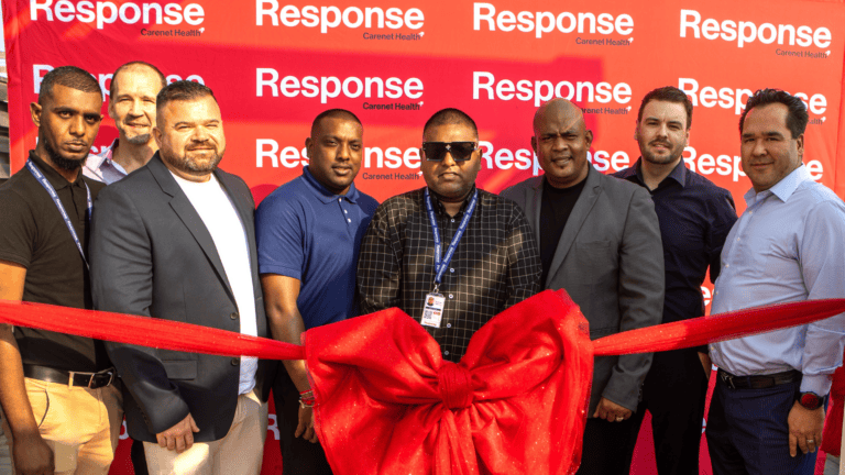 Group of seven people standing behind a large red ribbon and bow at a ribbon-cutting event, posing in front of a red “Response by Carenet Health” branded backdrop.