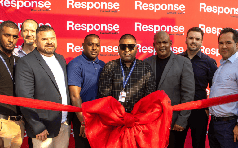 Group of seven people standing behind a large red ribbon and bow at a ribbon-cutting event, posing in front of a red “Response by Carenet Health” branded backdrop.